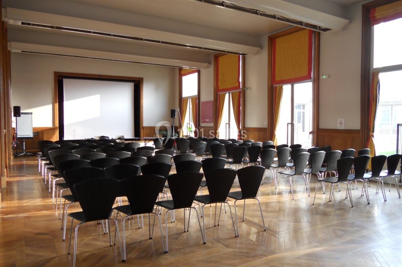Salle de conférence lumineuse avec des rangées de chaises noires face à un écran de projection.