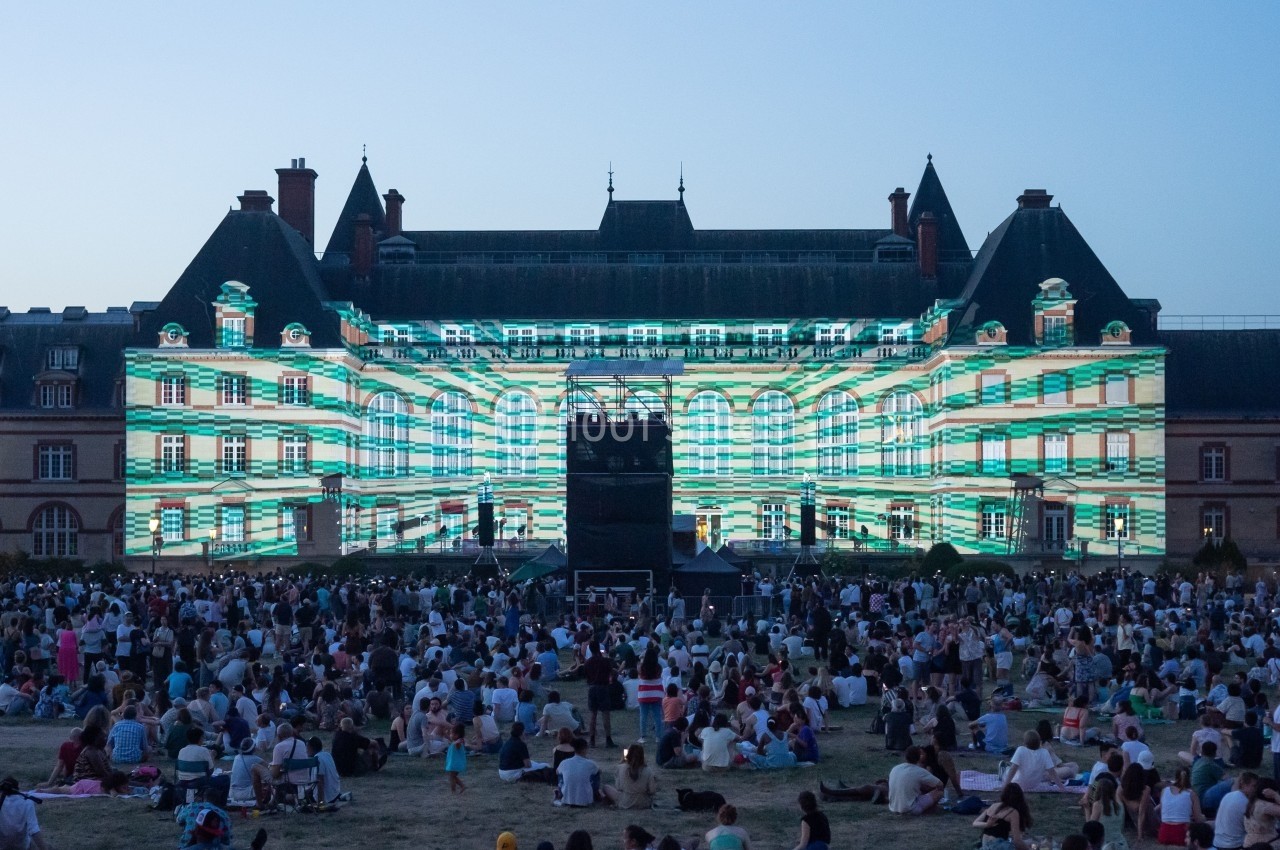 Foule assise devant un bâtiment historique illuminé par des projections lumineuses géométriques au crépuscule.