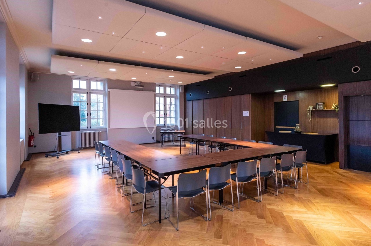 Salle de réunion lumineuse avec tables disposées en U, chaises, écran de projection et parquet en bois.