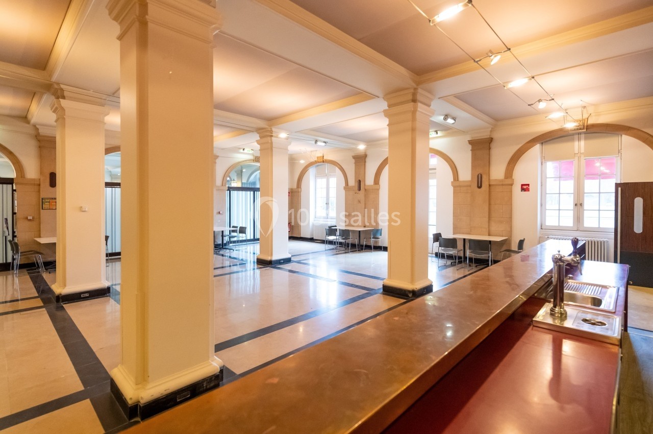 Salle lumineuse avec colonnes, sol en marbre, tables et chaises espacées, éclairage au plafond et grandes fenêtres.