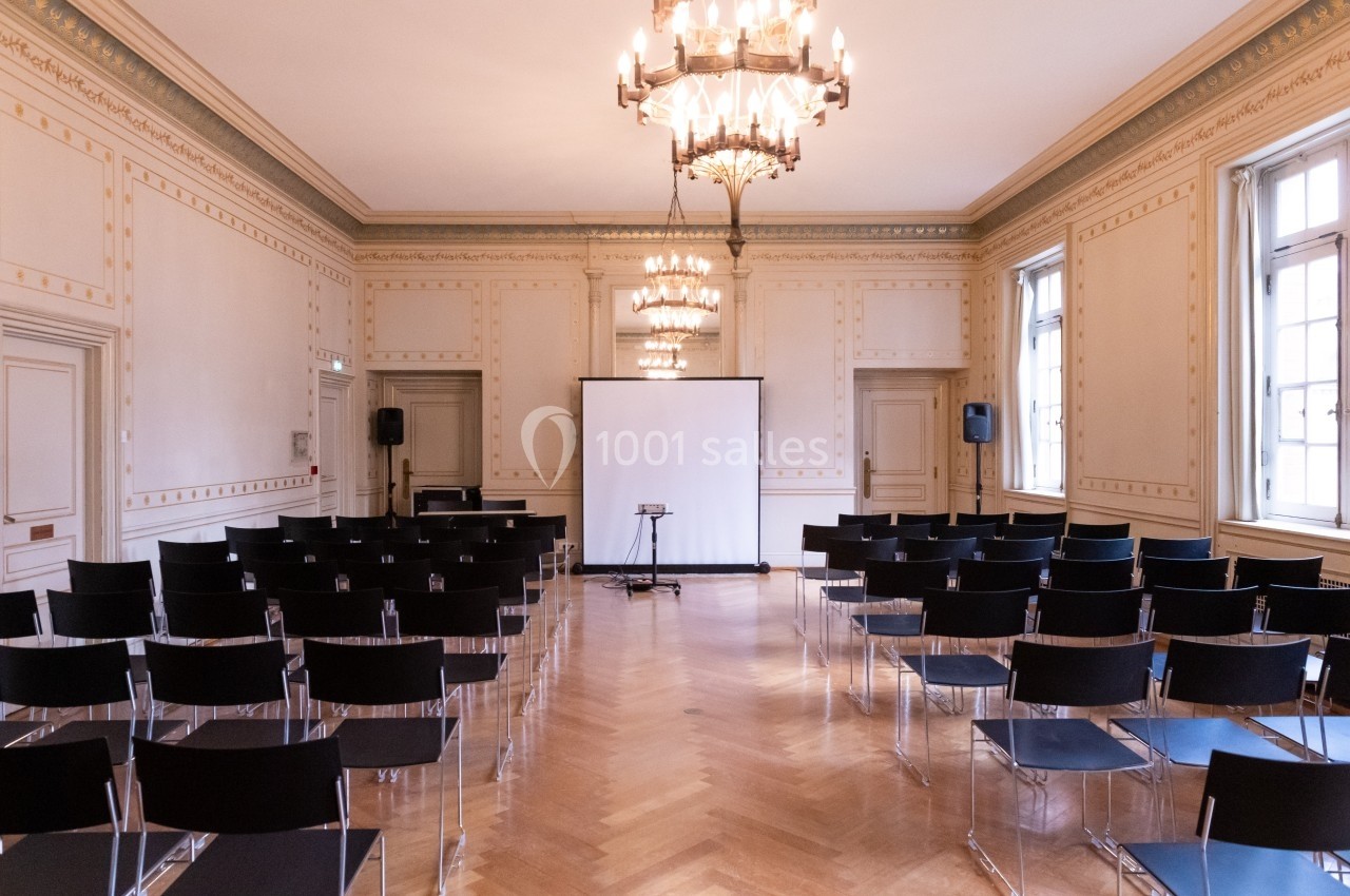 Salle de conférence lumineuse avec rangées de chaises noires, écran de projection et lustres suspendus.