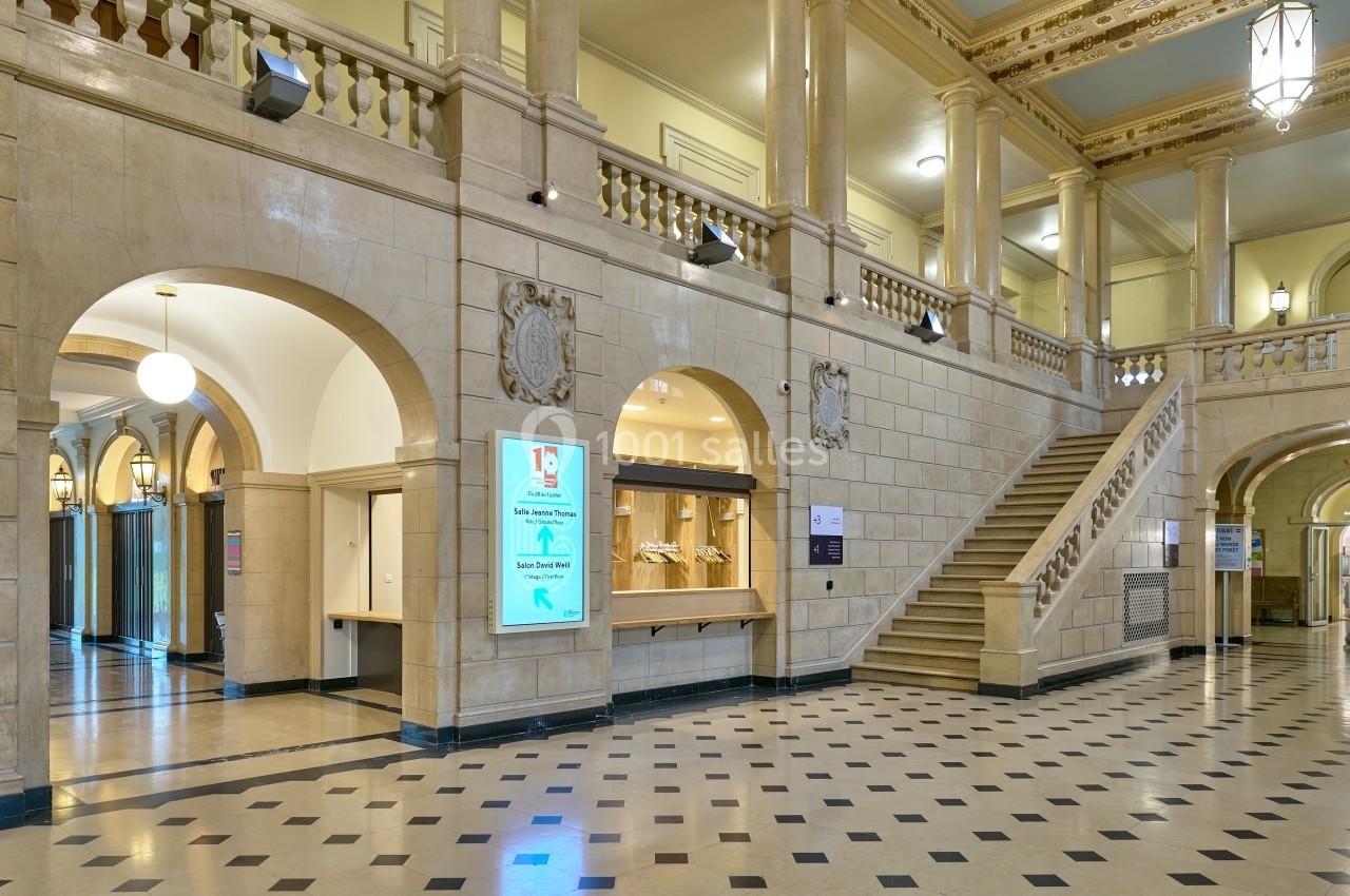 Hall spacieux avec sol en damier, escalier en pierre, arches, balustrades et panneaux d'information lumineux.