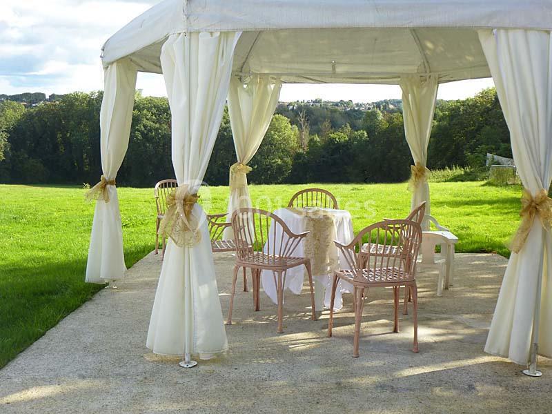 Tonnelle blanche avec rideaux ouverts, chaises et table décorées, située sur une terrasse en plein air avec vue sur un…