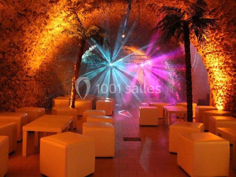 Salle voûtée avec éclairages colorés, palmiers décoratifs et mobilier blanc composé de tables et sièges cubiques.