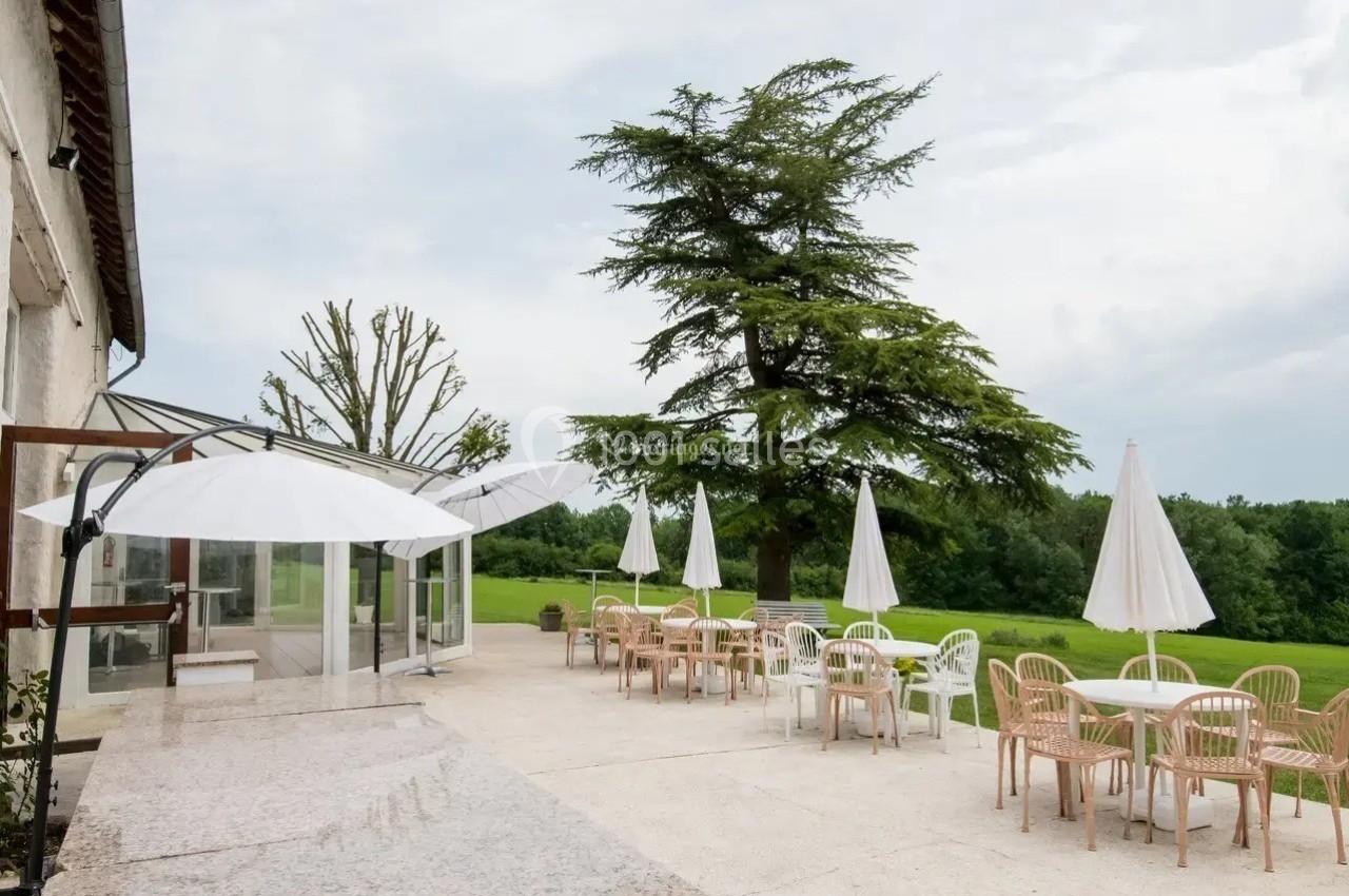 Terrasse avec tables, chaises et parasols blancs, entourée de verdure et d'arbres sous un ciel nuageux.