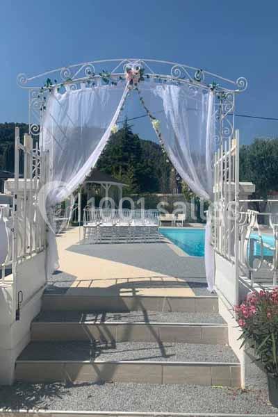 Arche décorée de voilages blancs et de fleurs, donnant sur une piscine entourée de chaises et d'arbres.