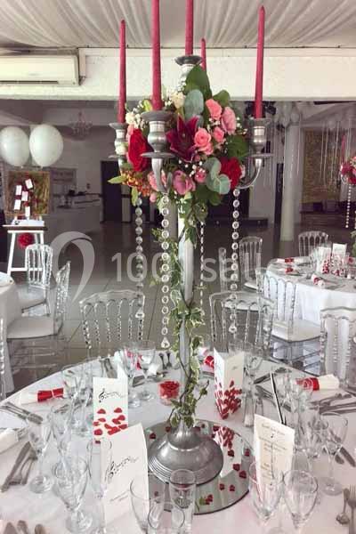 Centre de table avec chandelier argenté, bougies rouges, fleurs et perles, entouré de vaisselle sur une table dressée.