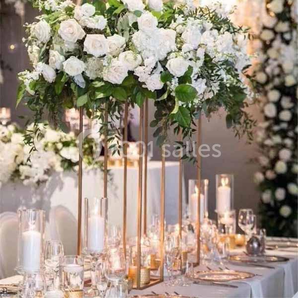 Centre de table floral avec des roses blanches et feuillages, sur une structure dorée, entouré de bougies.