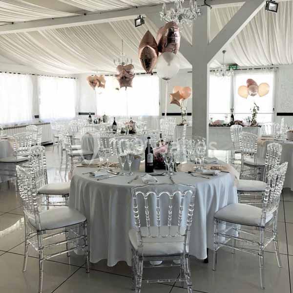 Salle de réception lumineuse décorée avec des ballons, des tables rondes dressées et des chaises transparentes.
