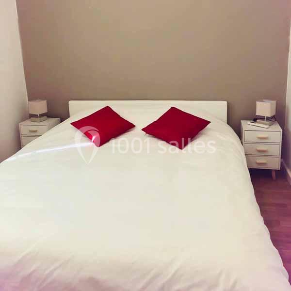 Lit double avec drap blanc, deux coussins rouges et tables de chevet blanches sur un sol en parquet.