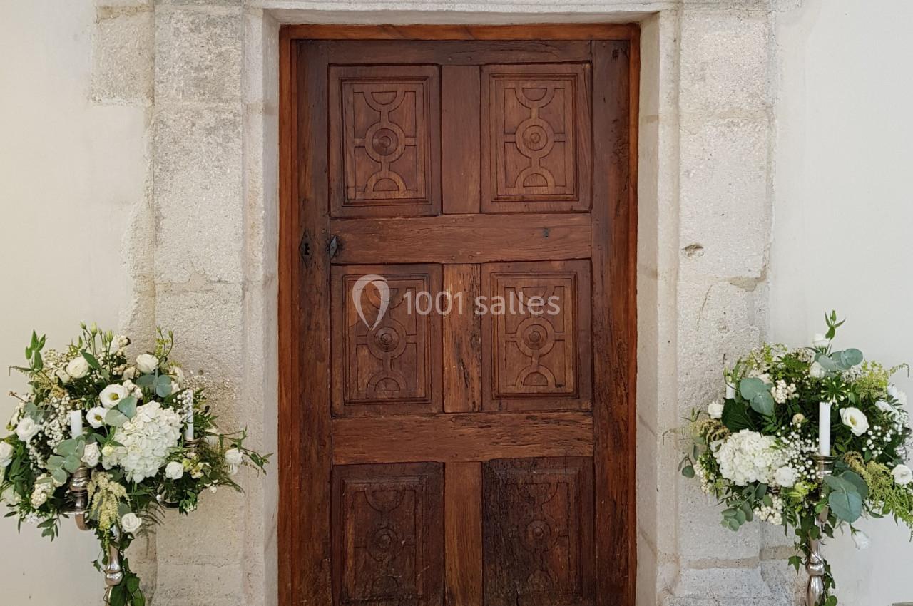Porte en bois sculptée encadrée de pierre, décorée de deux arrangements floraux sur des supports métalliques.