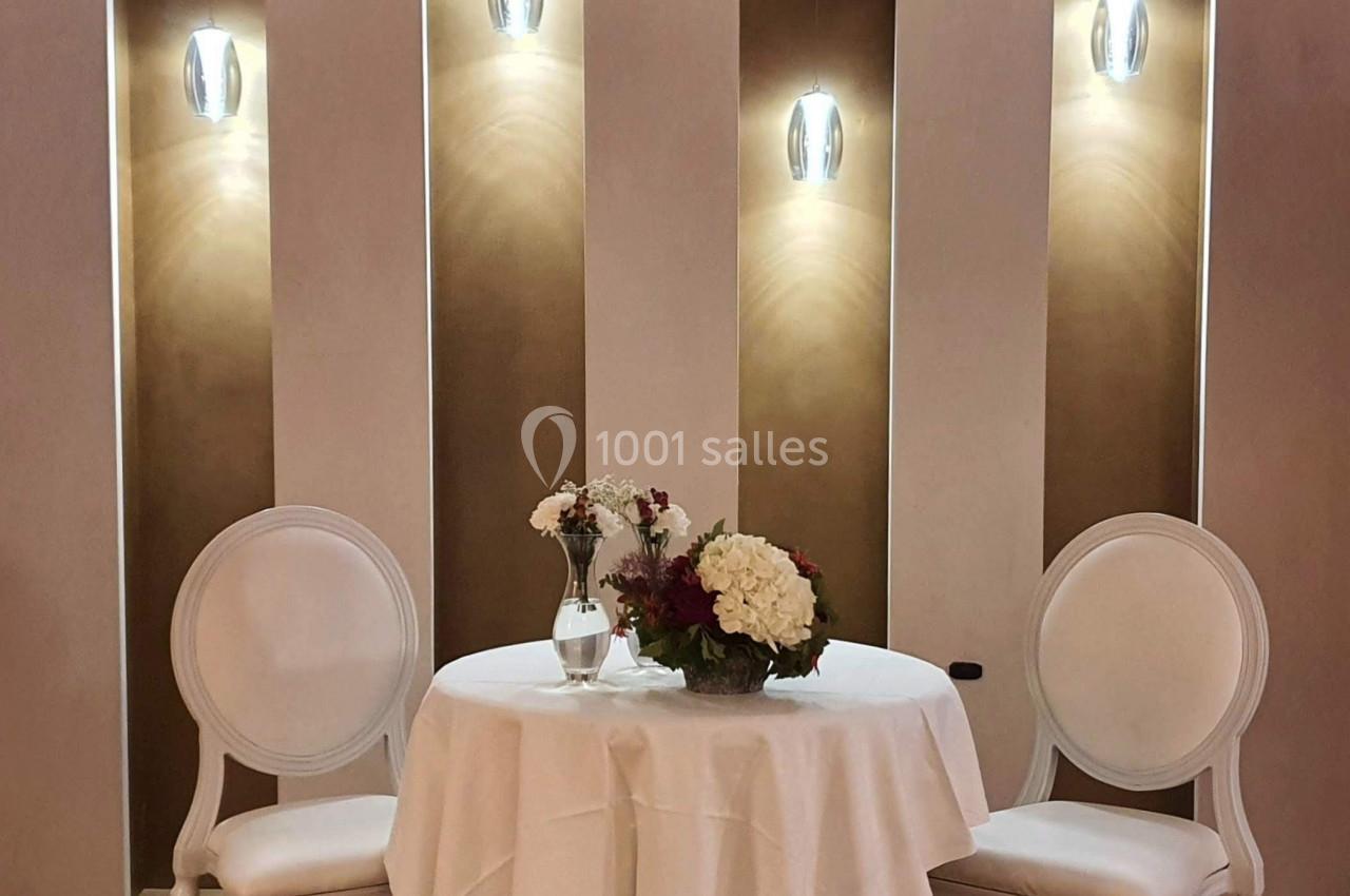 Table ronde avec nappe blanche, deux chaises blanches et un bouquet de fleurs, éclairée par des appliques murales.