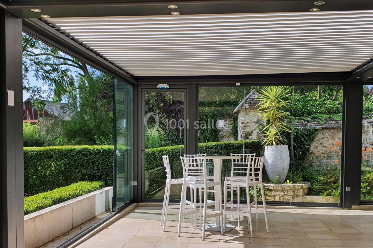 Terrasse couverte avec toit à lames orientables, mobilier blanc et vue sur un jardin verdoyant.