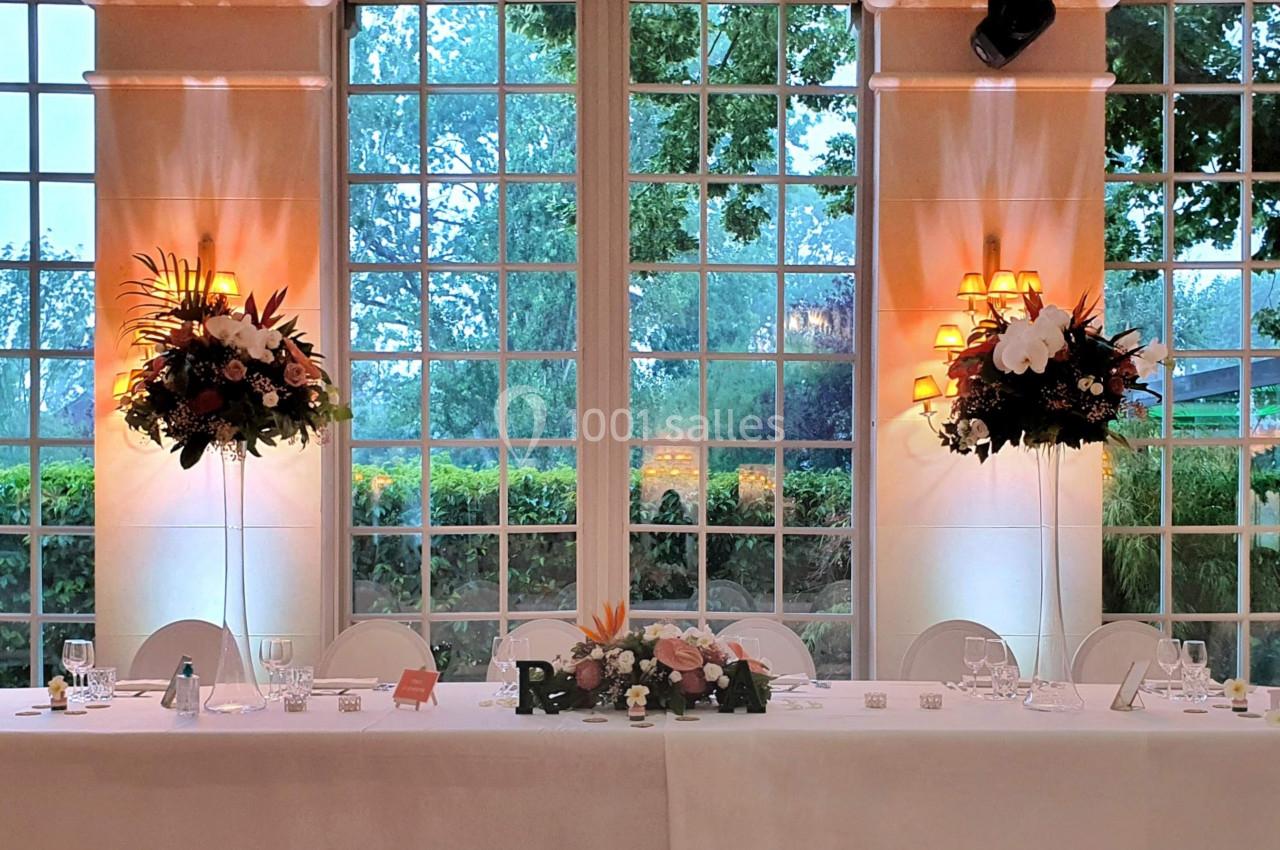 Salle de réception lumineuse avec une grande table décorée de fleurs, devant des baies vitrées donnant sur un jardin.
