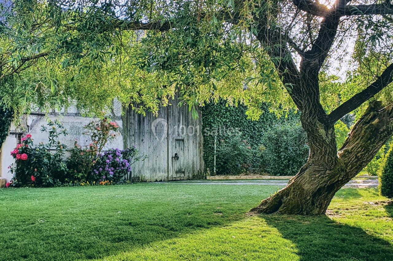 Jardin verdoyant avec un grand arbre, des fleurs colorées et un mur en pierre partiellement couvert de végétation.