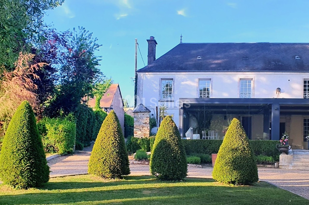 Jardin avec allée bordée de buis taillés, devant une grande maison blanche entourée de verdure.