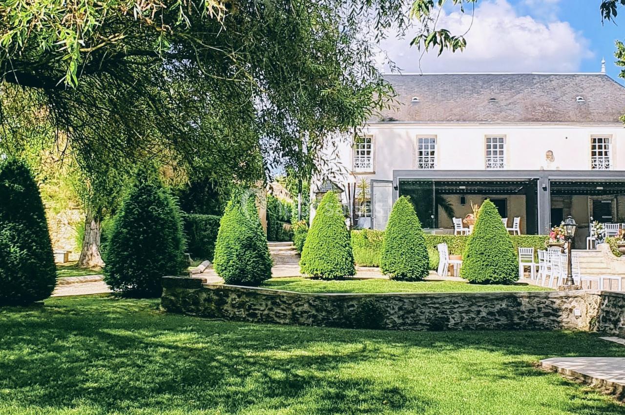 Jardin verdoyant avec pelouse, arbres et arbustes taillés, devant une maison blanche à toit d'ardoise.