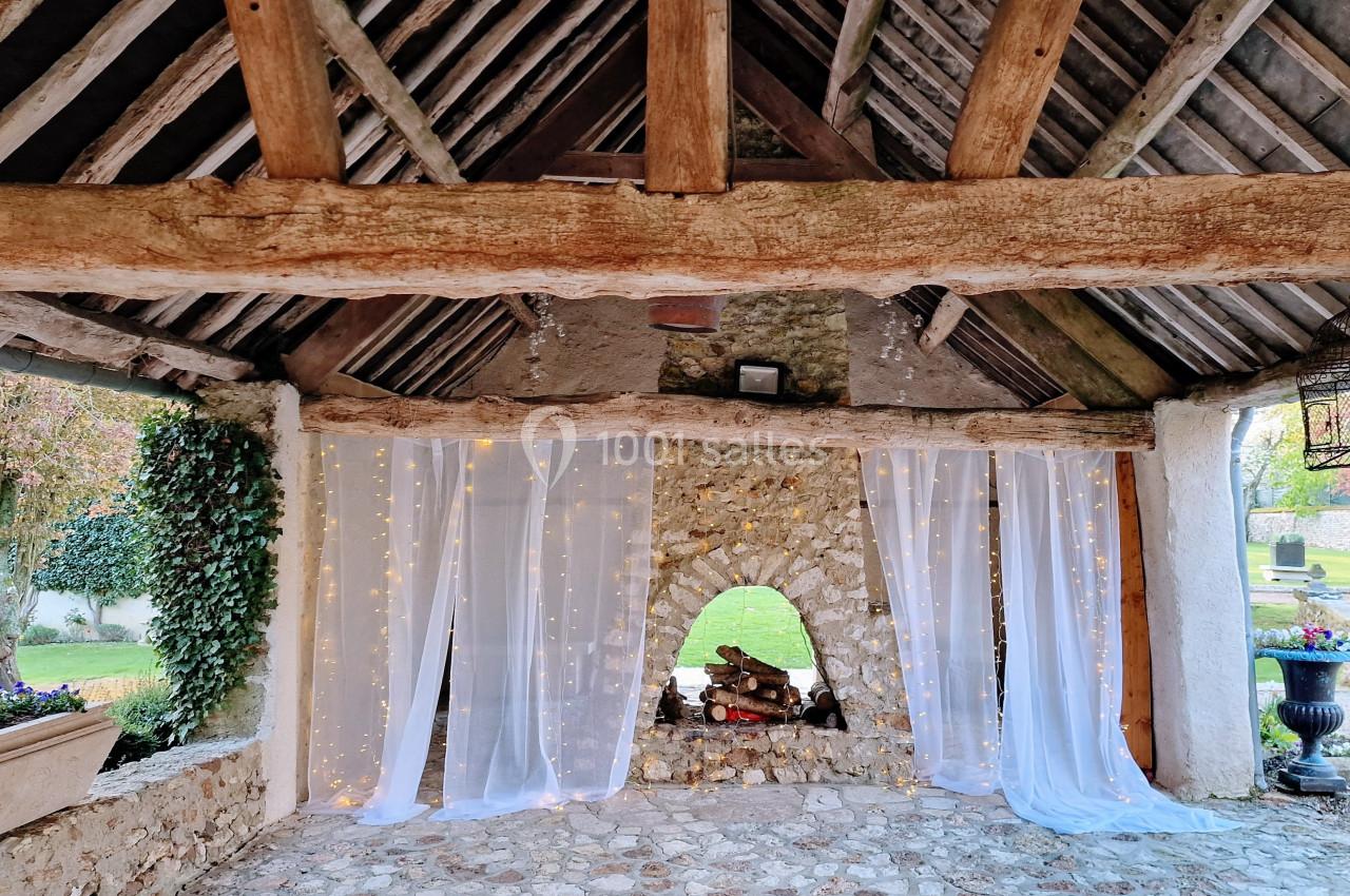 Structure en pierre avec toit en bois, rideaux blancs lumineux et ouverture centrale décorée de bûches.