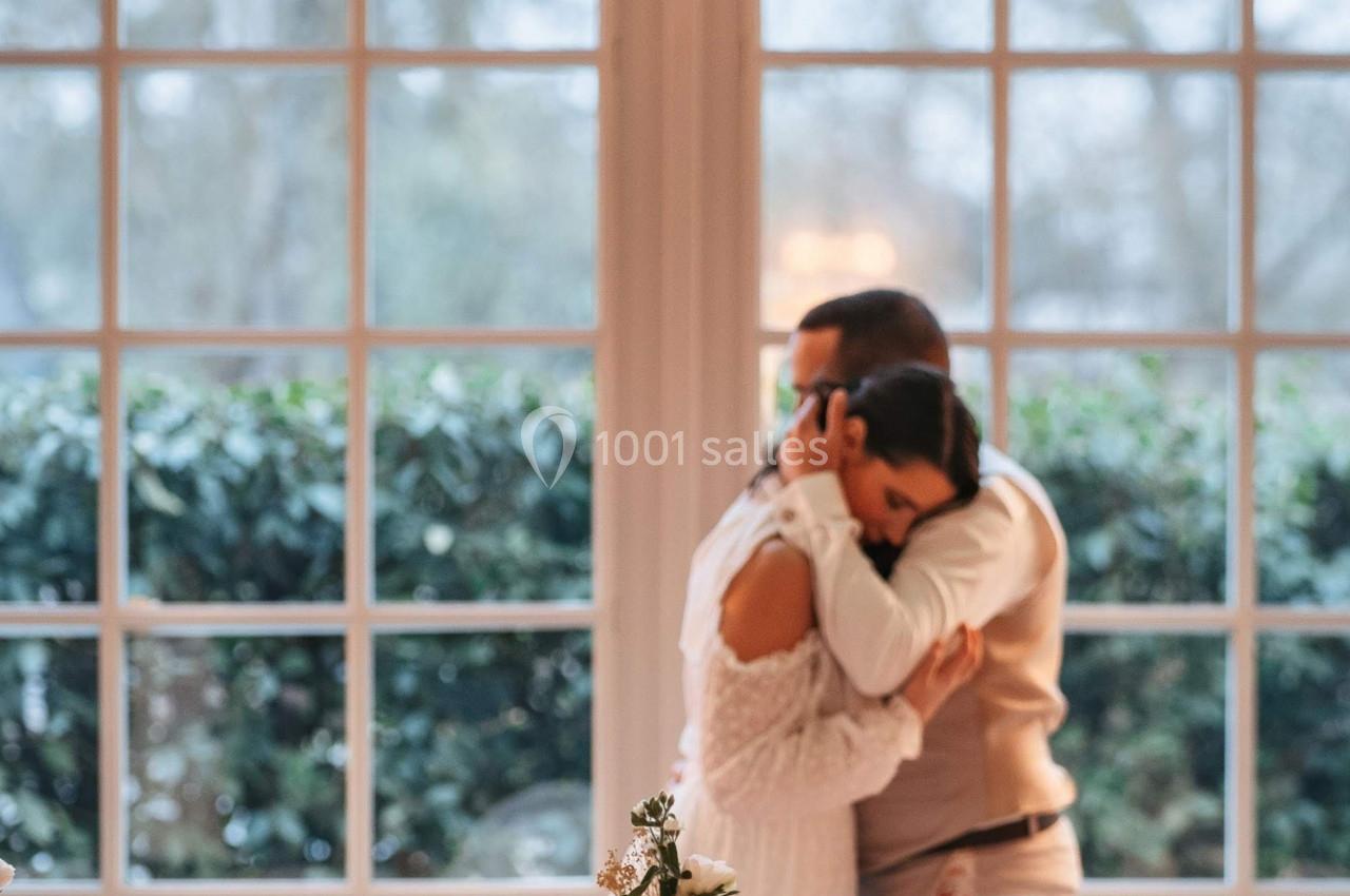 Un couple s'enlace devant une grande baie vitrée, avec une table décorée de fleurs et de feuillages au premier plan.