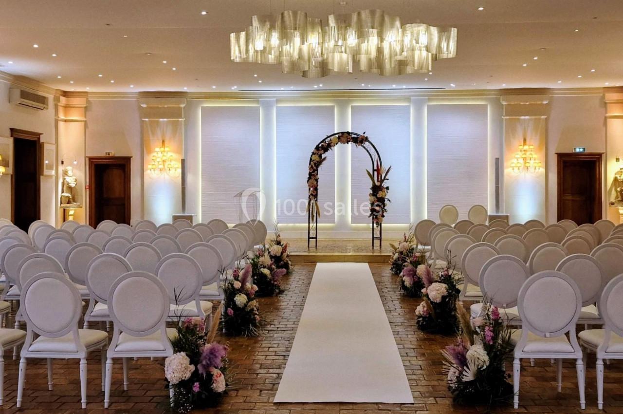 Salle de réception lumineuse aménagée pour une cérémonie, avec des rangées de chaises blanches et une arche fleurie au…
