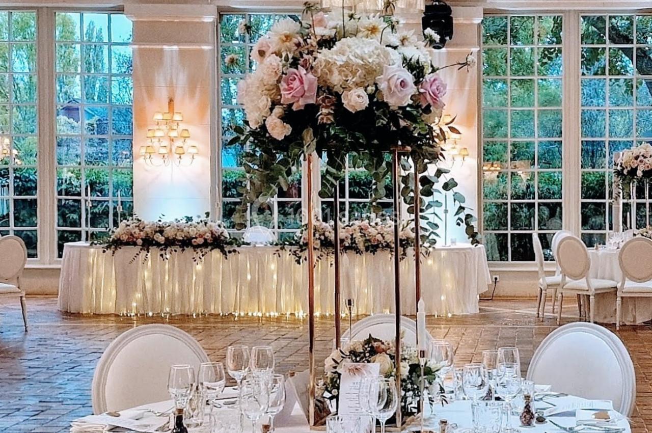 Salle de réception élégante avec tables décorées de fleurs, chaises blanches et grandes baies vitrées donnant sur un jardin.