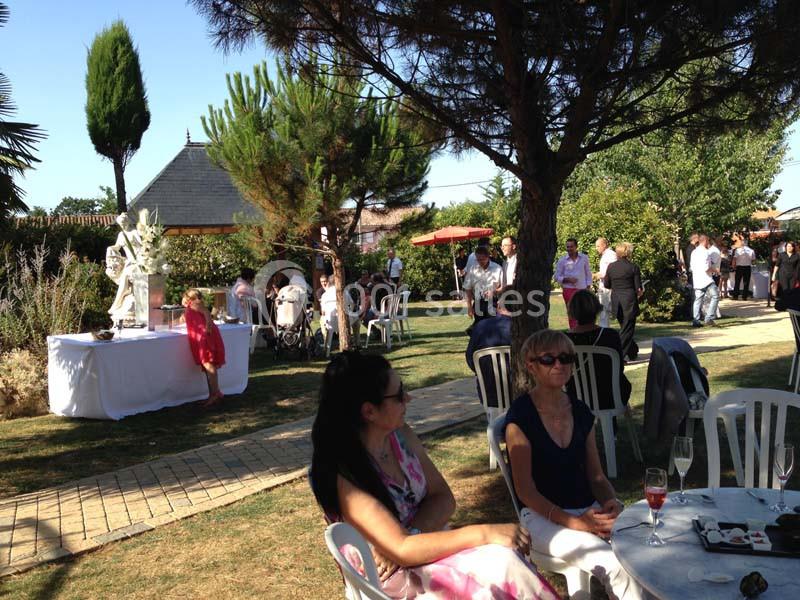 Des personnes assises et debout dans un jardin lors d'un événement en plein air, avec des tables et des arbres autour.