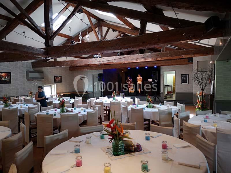 Salle de réception avec tables rondes décorées, poutres apparentes et une scène en arrière-plan.