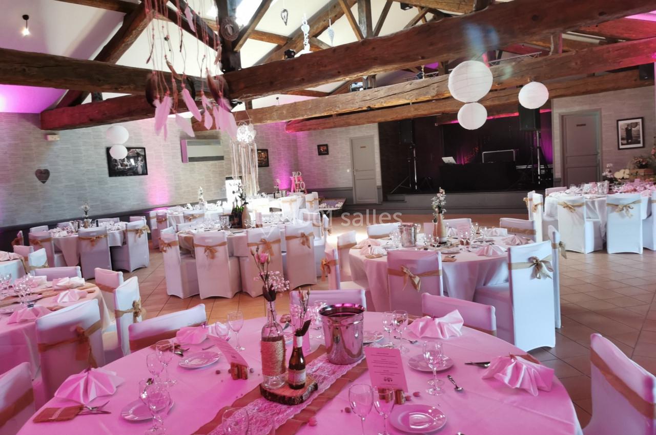 Salle de réception décorée pour un mariage avec tables dressées, nappes roses, chaises blanches et éclairage tamisé.