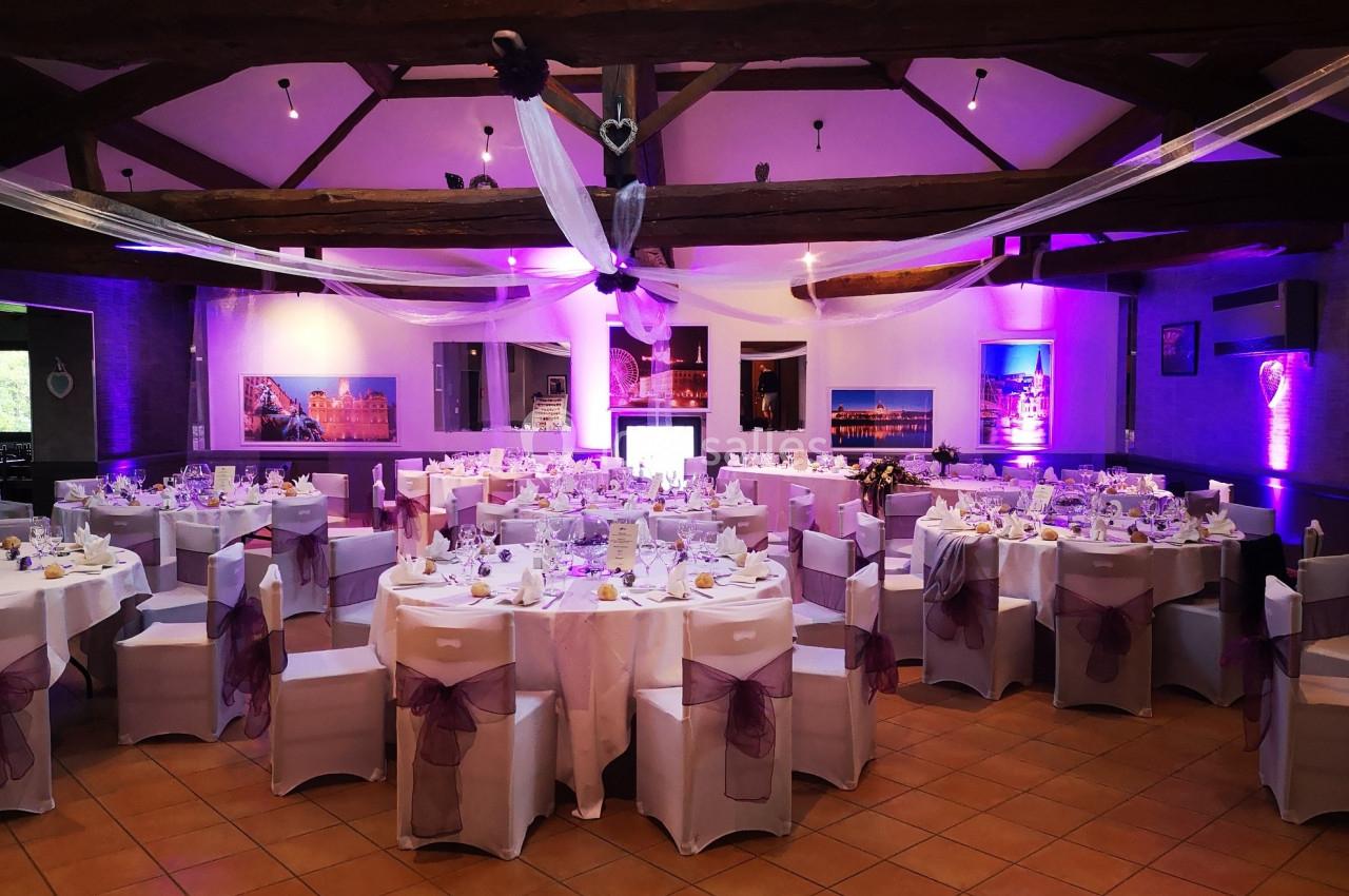 Salle de réception décorée avec des tables rondes dressées, nappes blanches et nœuds violets sur les chaises.