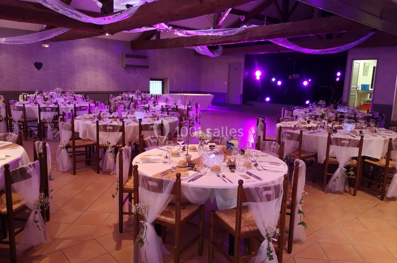 Salle de réception décorée pour un événement, avec tables rondes dressées, chaises ornées et éclairage violet.