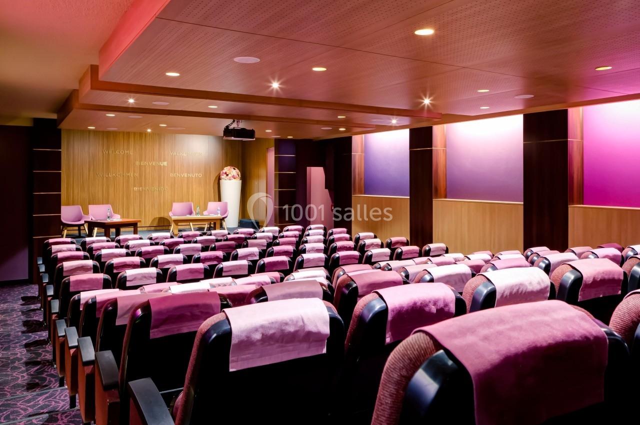 Salle de conférence moderne avec fauteuils rembourrés violets, scène en bois et éclairage tamisé.