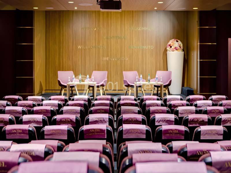 Salle de conférence avec rangées de sièges violets, une scène équipée de chaises et tables, et un décor en bois clair.
