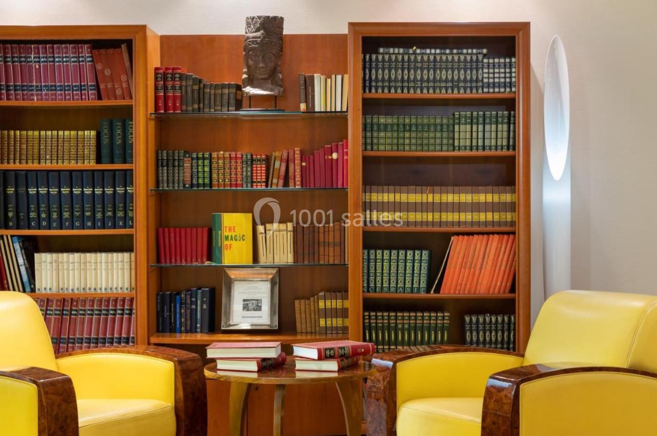 Bibliothèque avec étagères remplies de livres colorés, deux fauteuils jaunes et une table basse avec des livres ouverts.