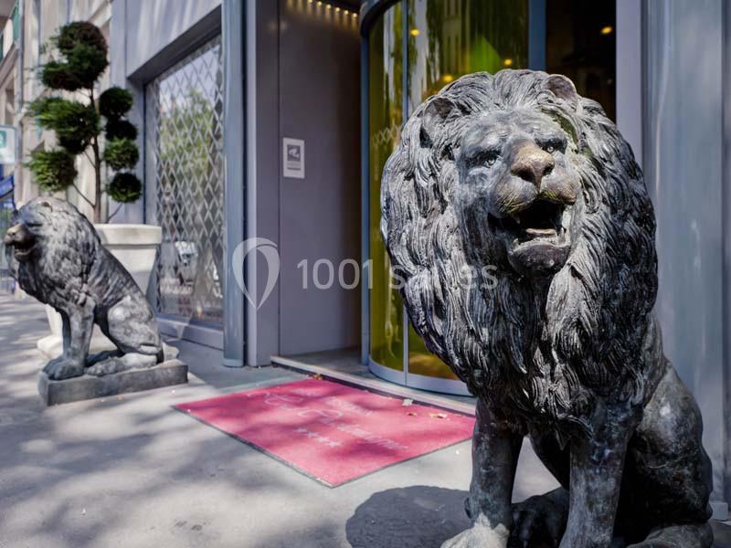 Deux statues de lions en métal encadrent l'entrée d'un bâtiment avec une porte vitrée et un tapis rouge.