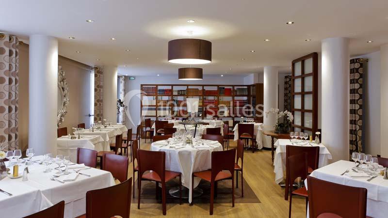 Salle de restaurant lumineuse avec tables dressées, chaises en bois, étagères de livres et décor sobre.