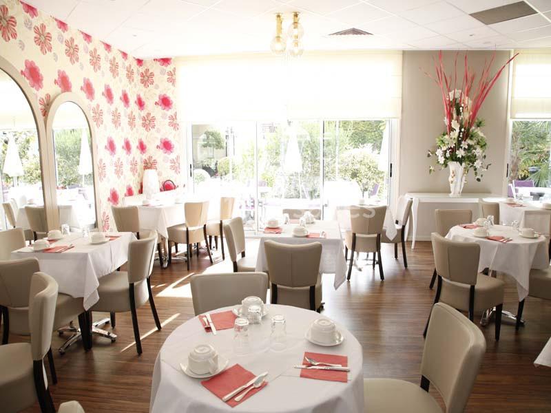 Salle de restaurant lumineuse avec tables dressées, nappes blanches, chaises beiges et décoration florale.