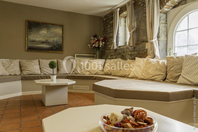 Salon chaleureux avec banquette d'angle beige, table basse blanche, mur en briques et fenêtre arrondie.