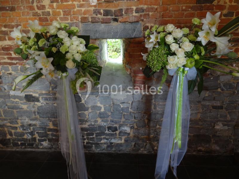Deux compositions florales blanches et vertes avec rubans, disposées devant un mur de briques et une petite ouverture.
