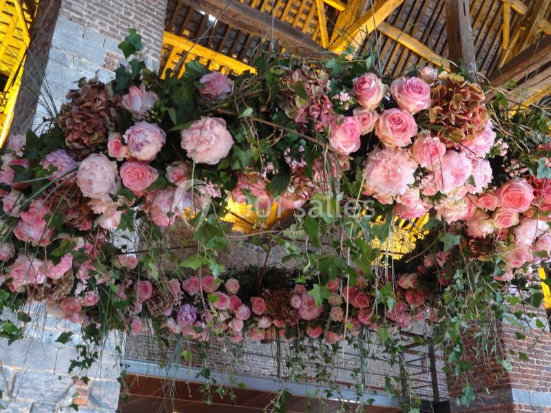 Guirlande suspendue de fleurs roses et feuillages dans un bâtiment en pierre et bois avec plafond en poutres apparentes.
