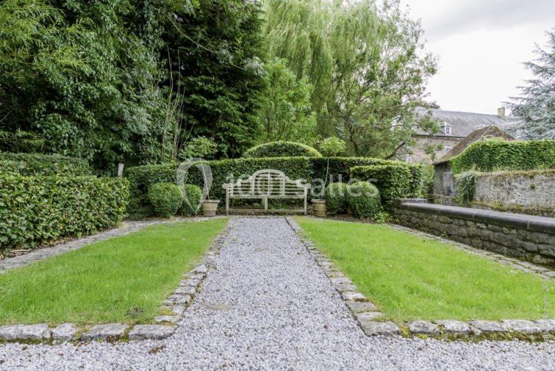 Allée de gravier bordée de pelouses et de haies, menant à un banc en bois blanc dans un jardin verdoyant.