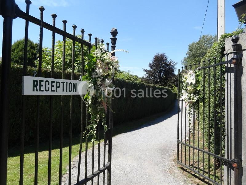 Portail en fer forgé ouvert, décoré de fleurs blanches, avec un panneau indiquant ’Réception’ et une allée en arrière-plan.