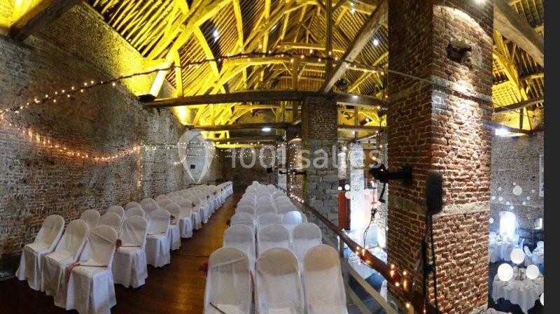 Salle en briques avec des poutres apparentes, décorée de guirlandes lumineuses et rangées de chaises blanches alignées.