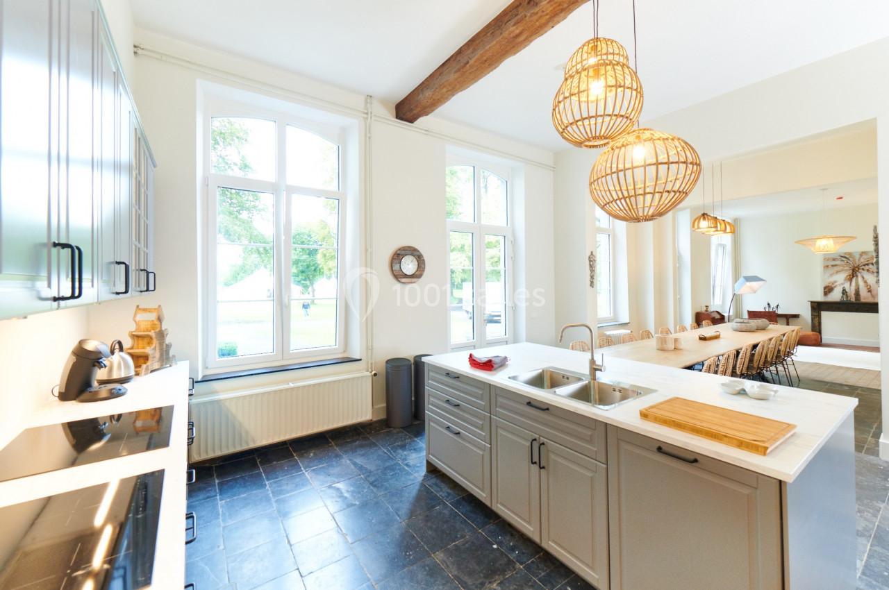 Cuisine lumineuse avec îlot central, grandes fenêtres, suspensions en rotin et sol en carrelage noir.