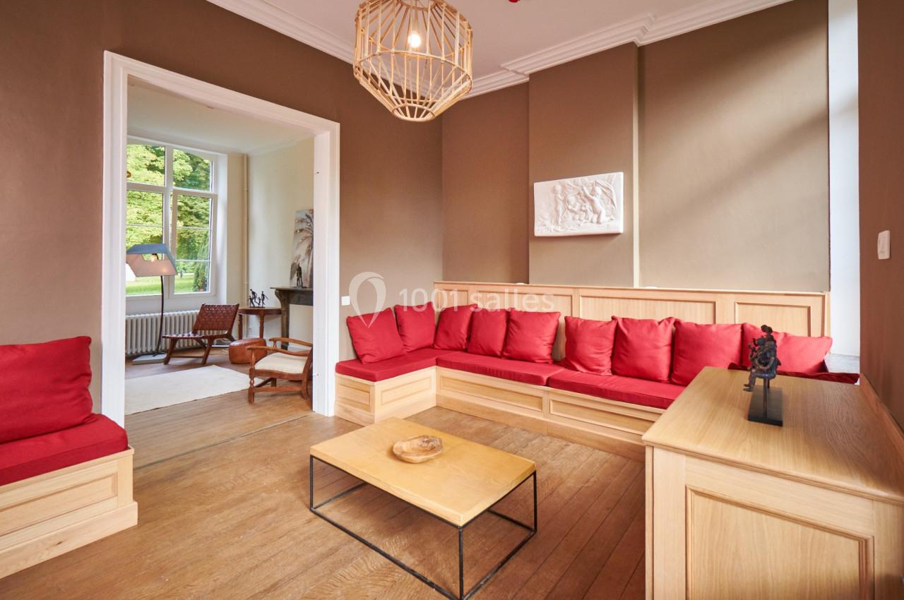 Salon avec canapé d'angle rouge, table basse en bois, parquet au sol et vue sur une pièce adjacente lumineuse.