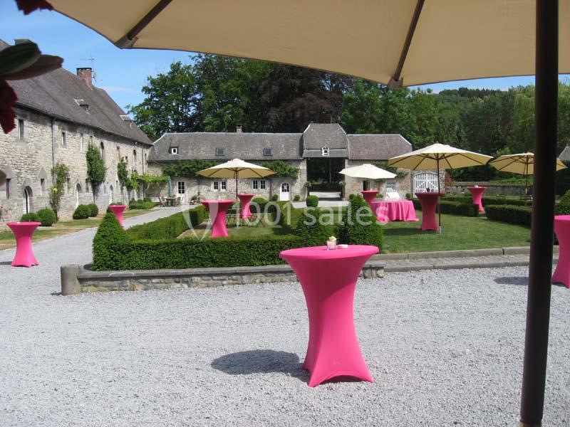 Cour extérieure d'un domaine avec des tables hautes roses et des parasols beiges, entourée de verdure et de bâtiments en…