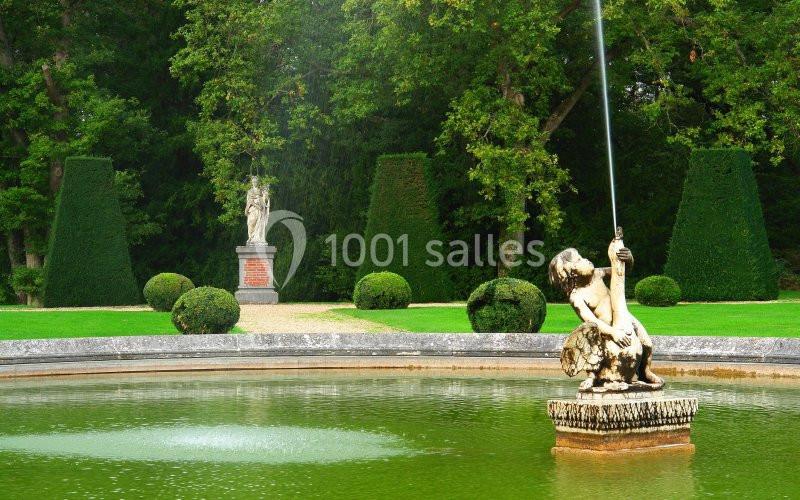 Statue d'enfant tenant un jet d'eau au centre d'un bassin, entourée de buissons taillés et d'arbres dans un jardin.
