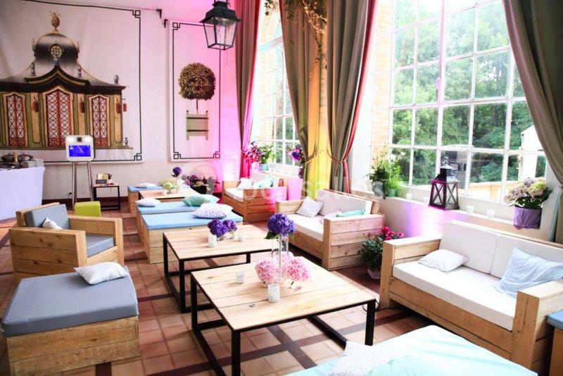 Salon lumineux avec mobilier en bois, coussins colorés, fleurs décoratives et grandes fenêtres donnant sur un jardin.