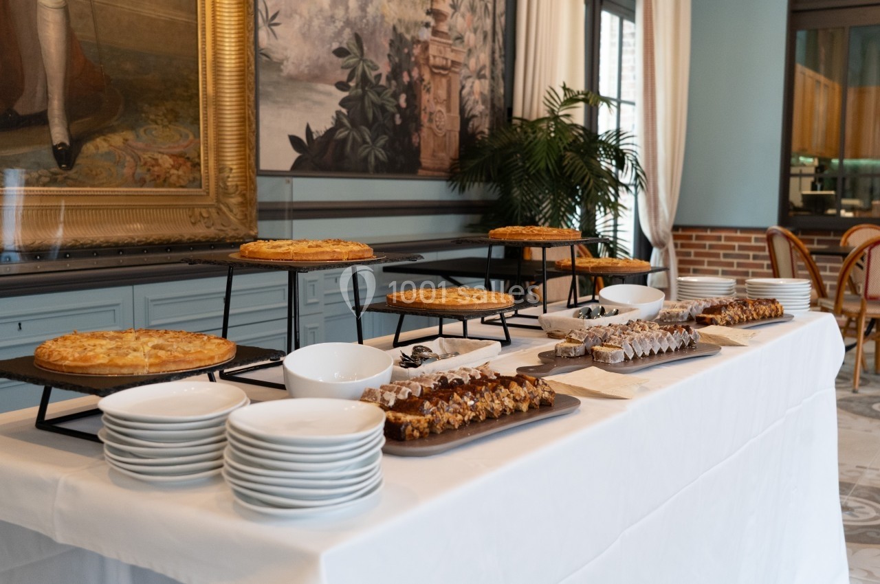 Table dressée avec des assiettes, des plats de tartes et de desserts variés, dans une salle décorée de tableaux et de…