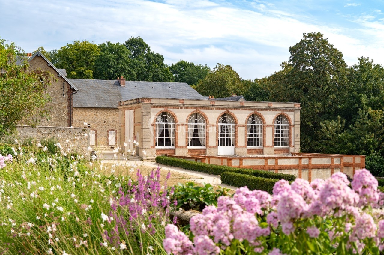 Bâtiment ancien avec grandes fenêtres en arc, entouré de verdure et de fleurs roses au premier plan.
