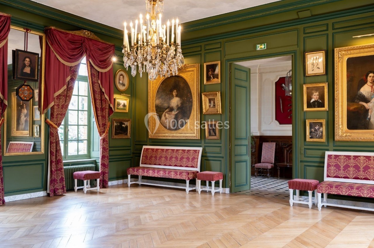 Salle élégante avec murs verts, tableaux encadrés, lustre doré, rideaux rouges et mobilier assorti.